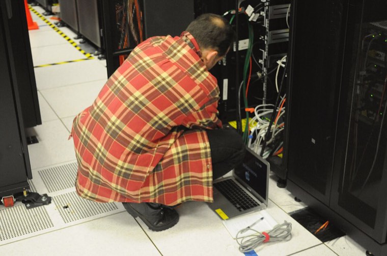Technicien accroupi devant une baie de serveurs, un ordinateur portable posé au sol, dans une salle informatique